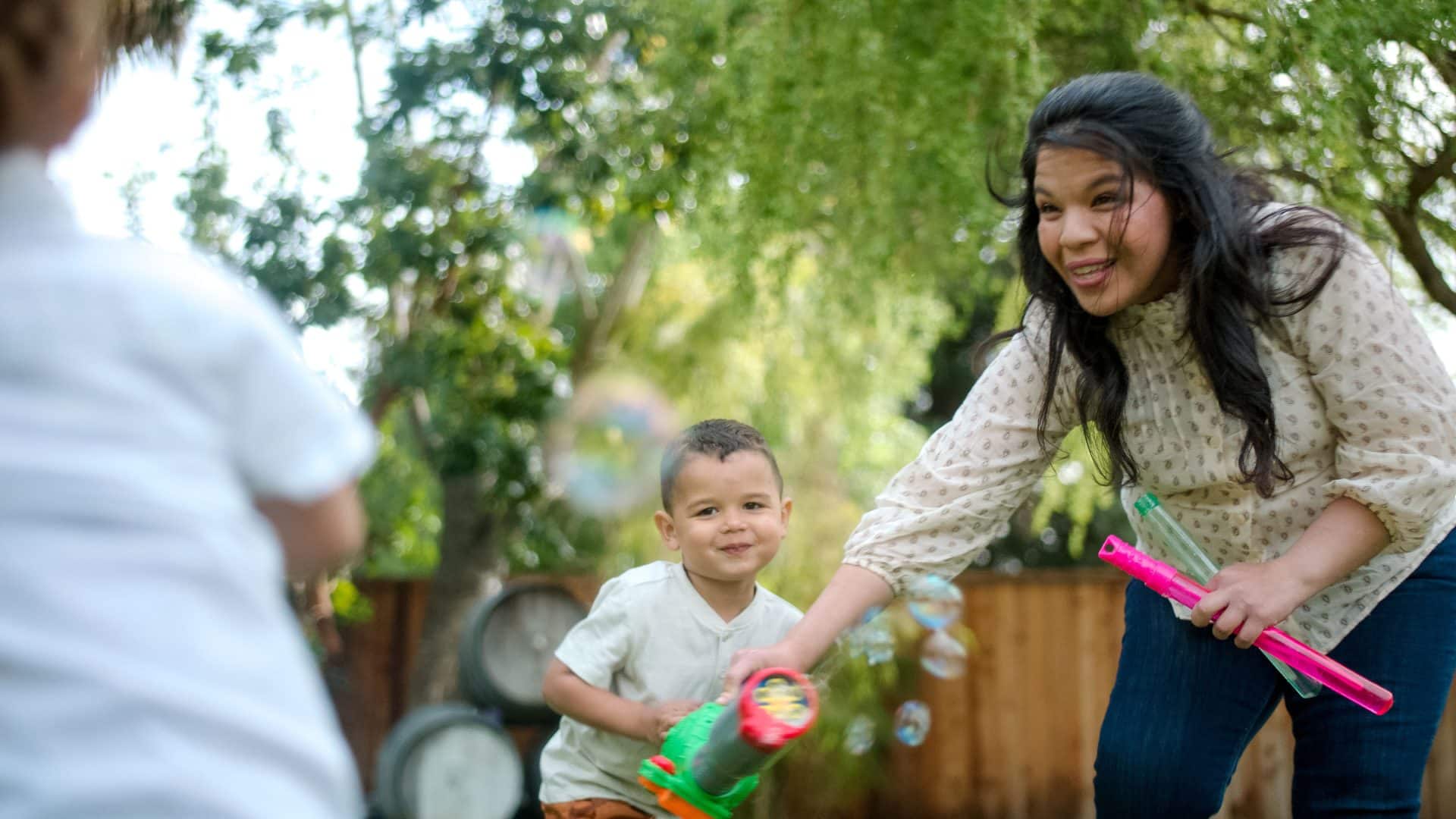 A homeschool nanny joins a woman and two children as they play with toy bubble guns outdoors, surrounded by trees.