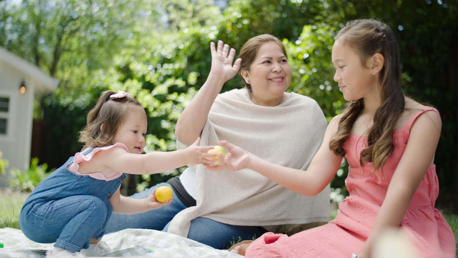 A woman and two young girls enjoy a picnic outdoors, smiling and playing with small yellow balls.