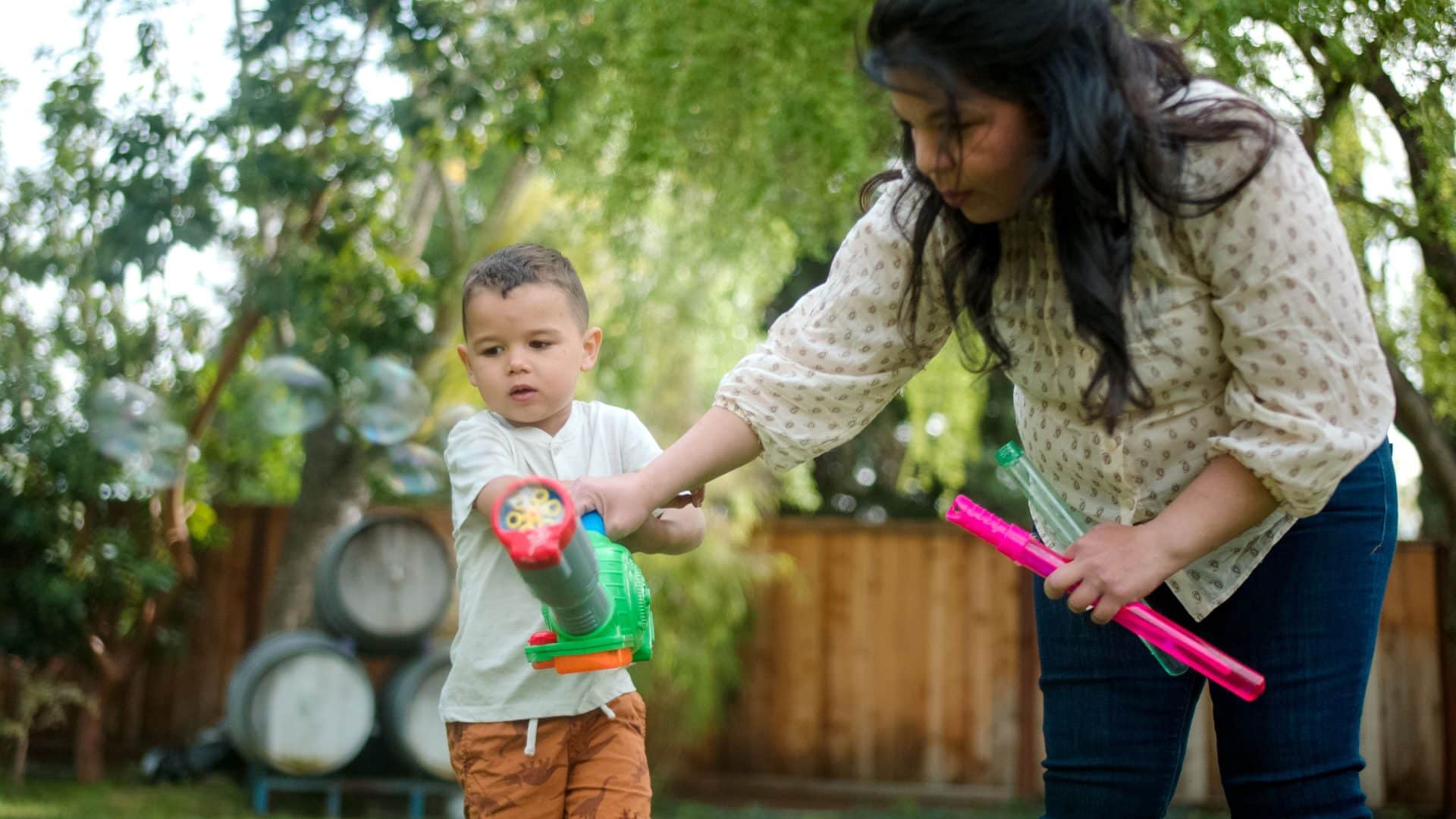 A woman and child play with bubble guns in a sunny backyard, surrounded by greenery and bubbles.