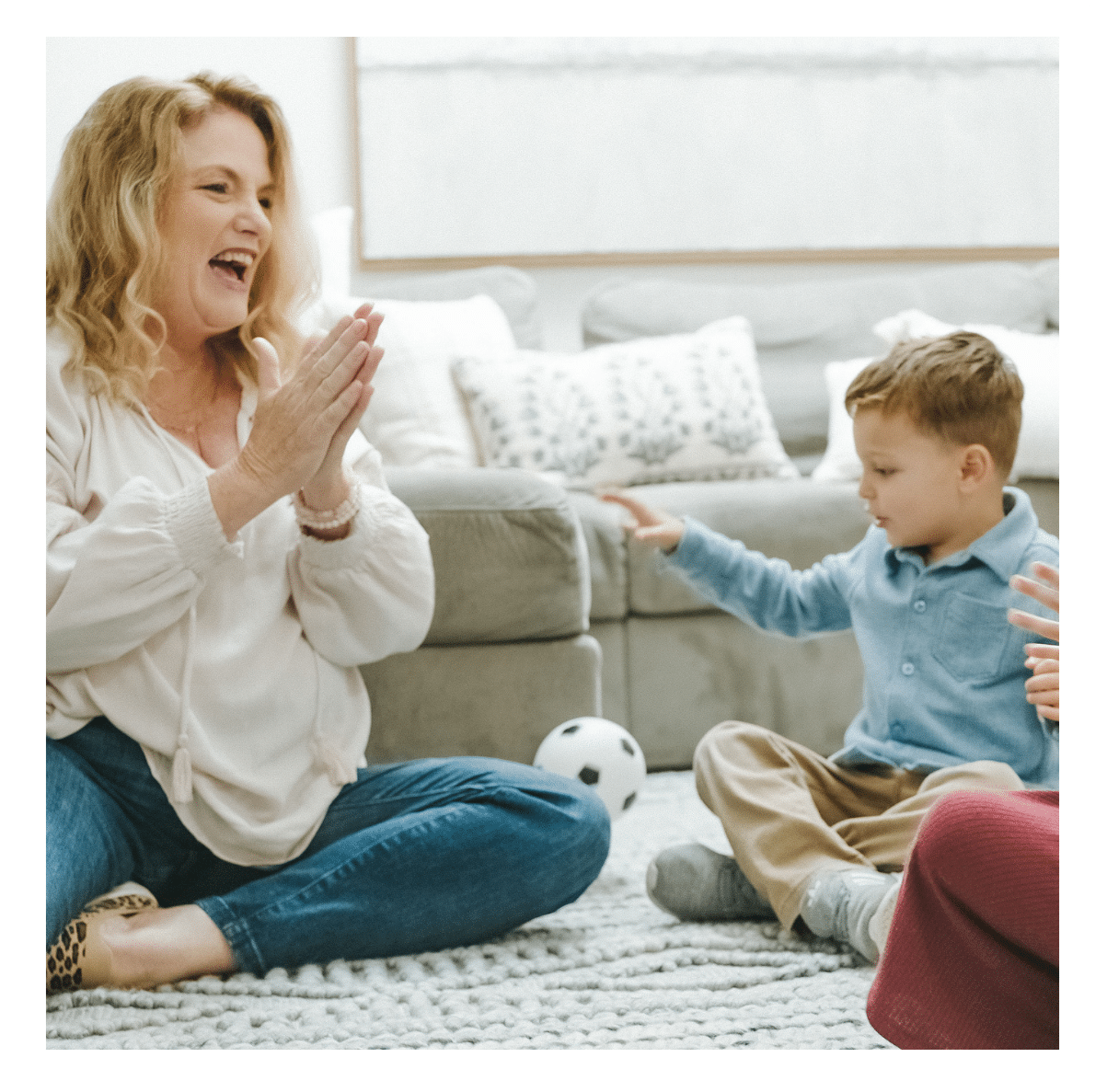 Woman clapping and smiling while sitting on the floor with a young boy playing with a ball indoors.