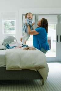 A woman lifts a smiling toddler above a bed, next to a laundry basket with clothes in a bright room.