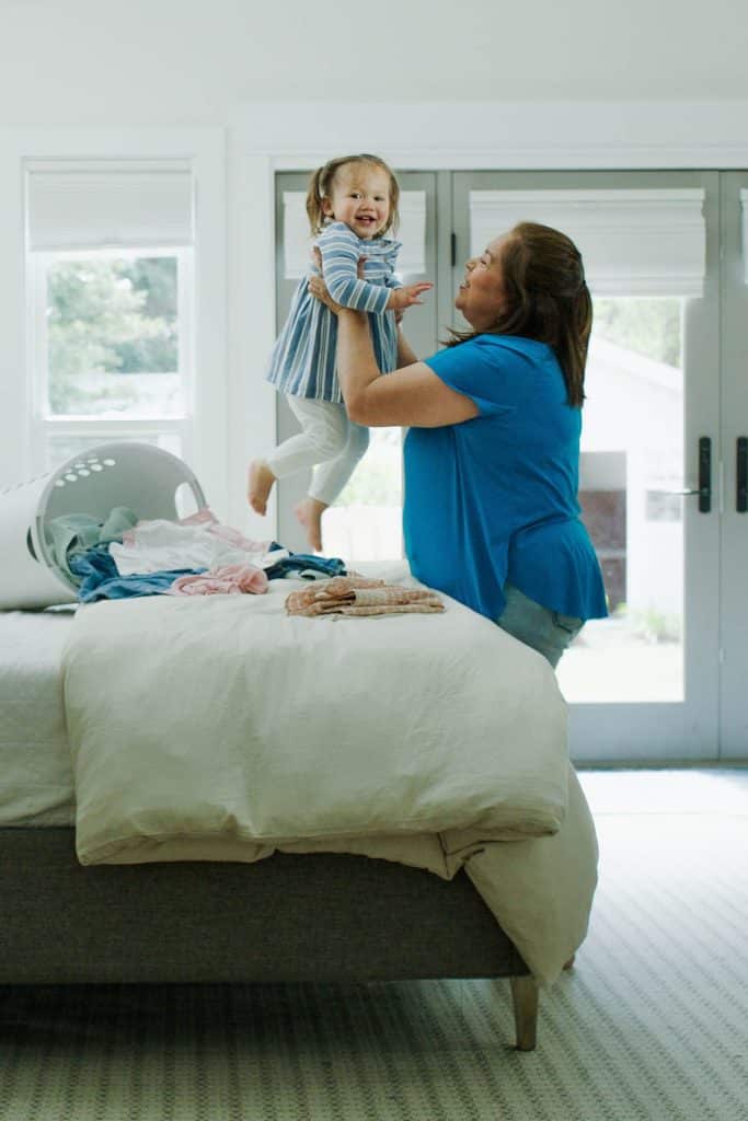 A woman lifts a smiling toddler above a bed, next to a laundry basket with clothes in a bright room.