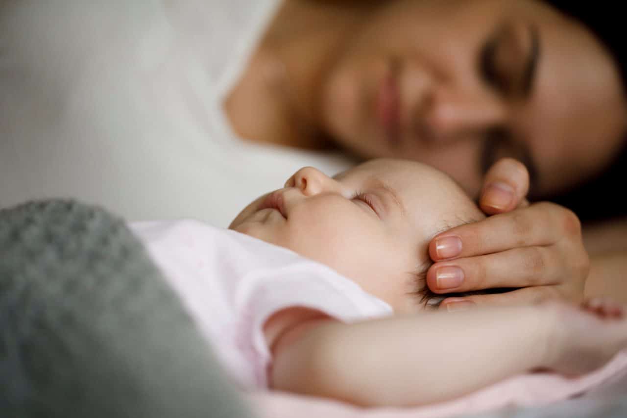 Baby sleeping peacefully, head gently caressed by a hand, lying next to a person resting in the background.