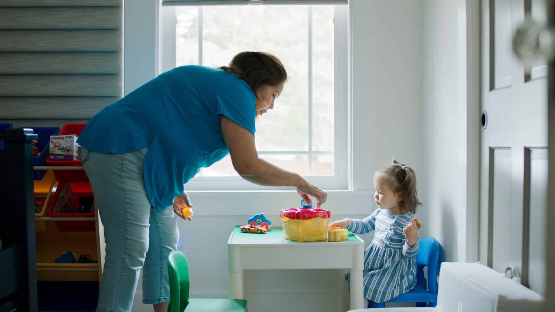 An adult and child engage in playful learning at a small table with toys near a window in a bright room, creating an ideal homeschool environment.