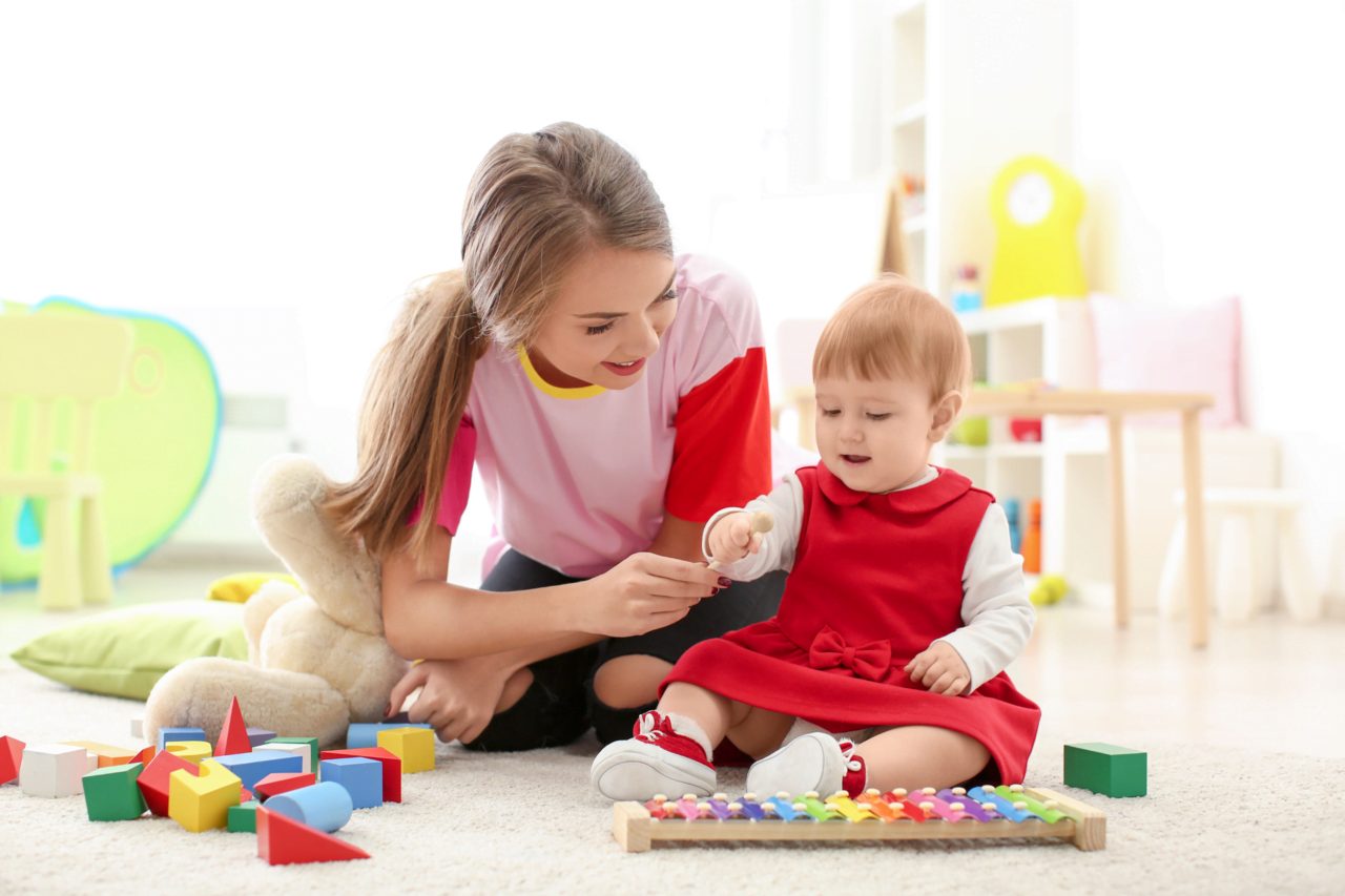 In a bright room with soft furnishings, a homeschool nanny engages a woman and toddler in playful learning, using colorful toys like a vibrant xylophone.