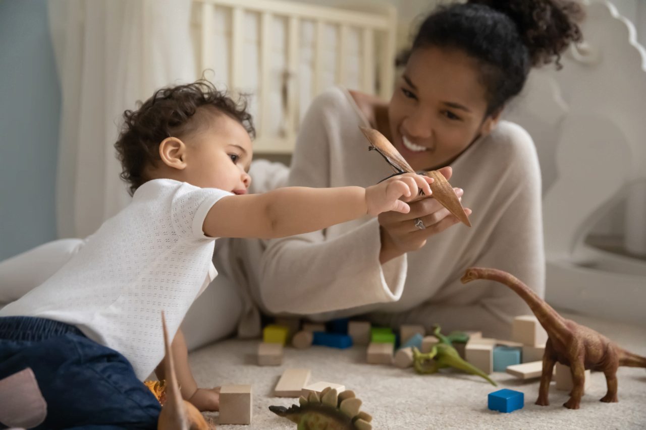 A mother and child, guided by their devoted homeschool nanny, play with toy dinosaurs and blocks on the floor, smiling and engaged.