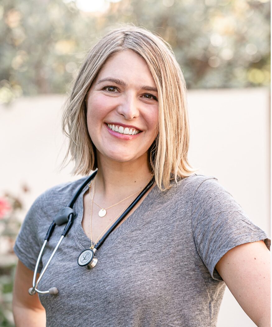 Smiling woman in a gray shirt with a stethoscope around her neck, standing outdoors.