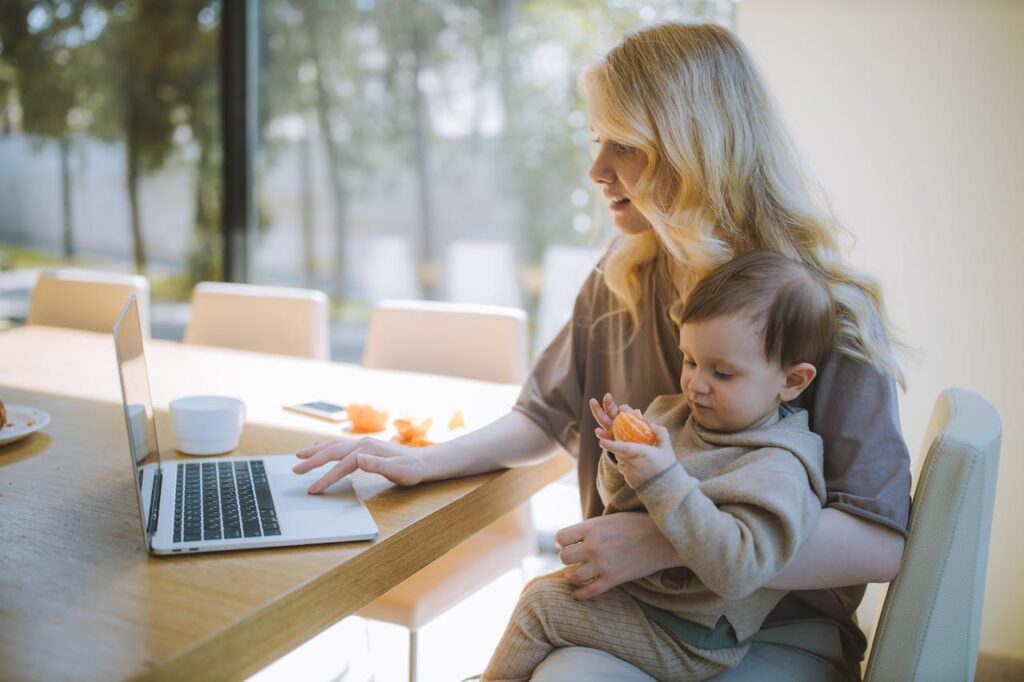 Woman working on a laptop at home while holding a baby on her lap, with a bright window in the background.