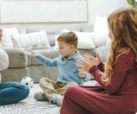 Children sitting on a carpeted floor, clapping and playing with a small soccer ball in a cozy living room.