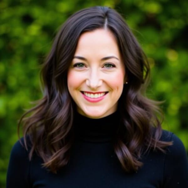 Person with long brown hair smiling, wearing a black top, standing in front of a blurred green background.