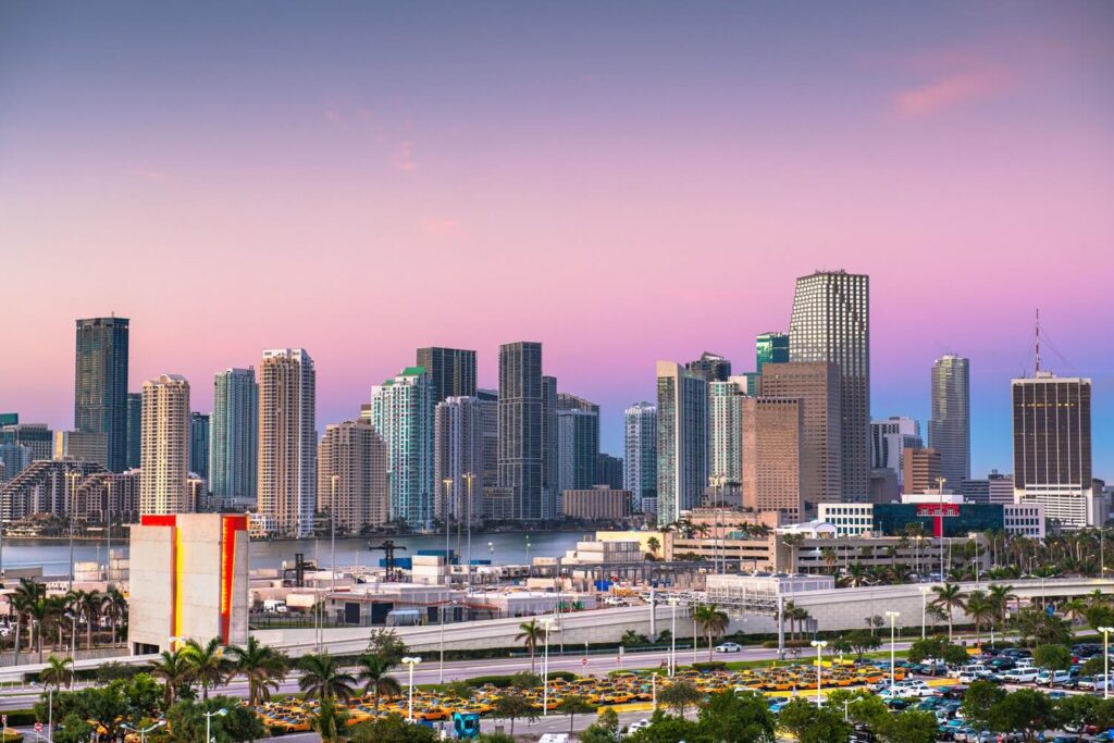 Miami skyline at sunset, featuring tall skyscrapers and a pastel-colored sky with palm trees in the foreground.