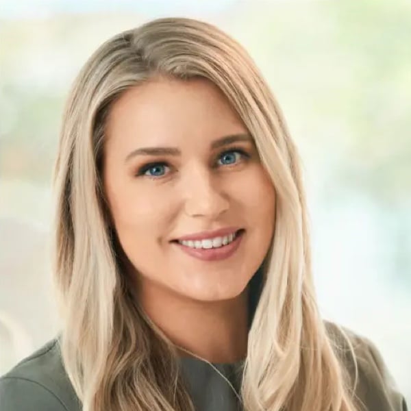 Smiling woman with long blonde hair and blue eyes, wearing a gray top against a softly blurred background.