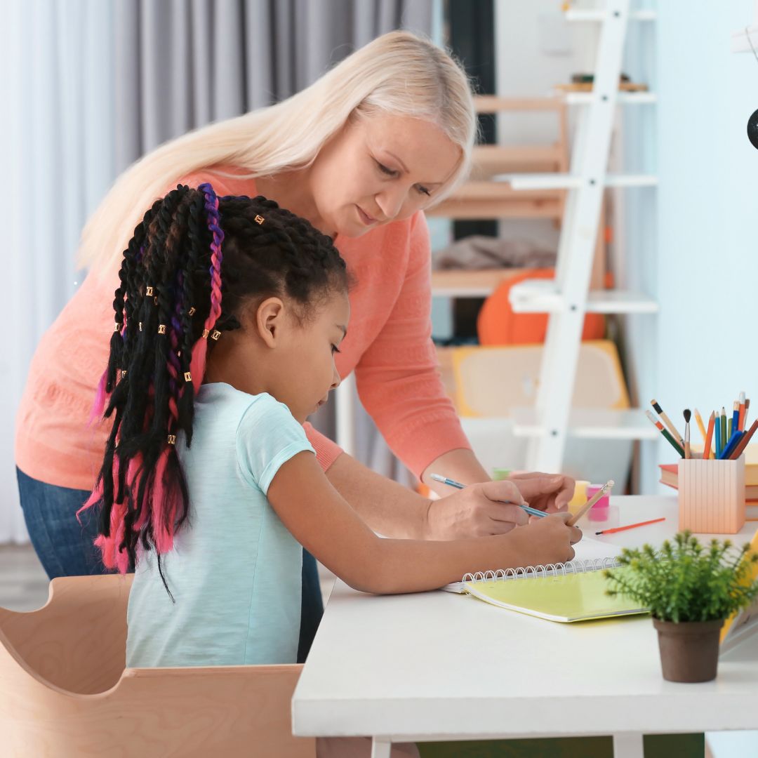 An educated nanny assists a girl with her homework at a white table, surrounded by vibrant pencils and a lush plant.