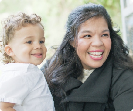 A woman and a toddler smile joyfully outdoors, with greenery in the background.