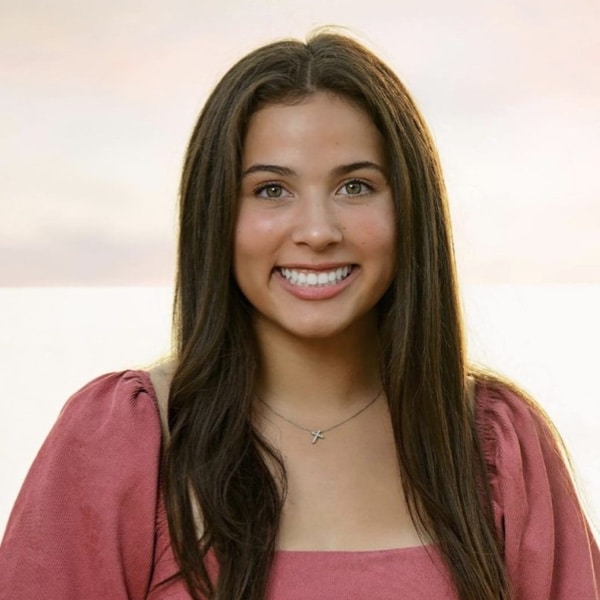 A woman with long dark hair, wearing a pink top, smiles against a blurred outdoor background.