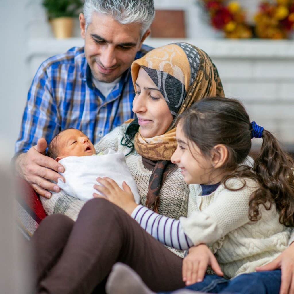 A family sits together, smiling warmly at their newborn wrapped in a white blanket, discussing infant sleep training. The cozy home setting is filled with warm colors, creating a perfect backdrop for this tender moment.