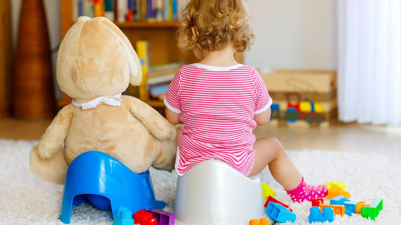 A child in red stripes and a teddy bear sit on potties, surrounded by colorful toy blocks on the carpet—a delightful scene capturing the essence of potty training.