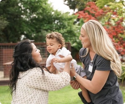 Two women smiling at a toddler held between them in a garden with trees in the background.