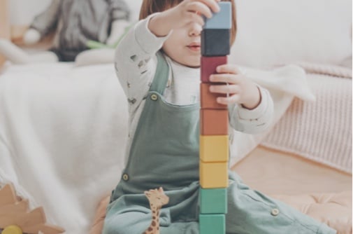 Child stacking colorful blocks, wearing green overalls, sitting on floor with a toy giraffe nearby.