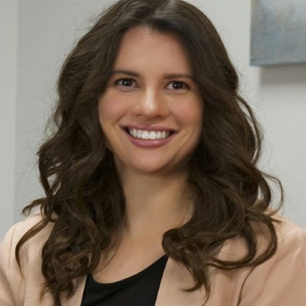 Smiling person with long, wavy dark hair wearing a light-colored jacket over a black top.