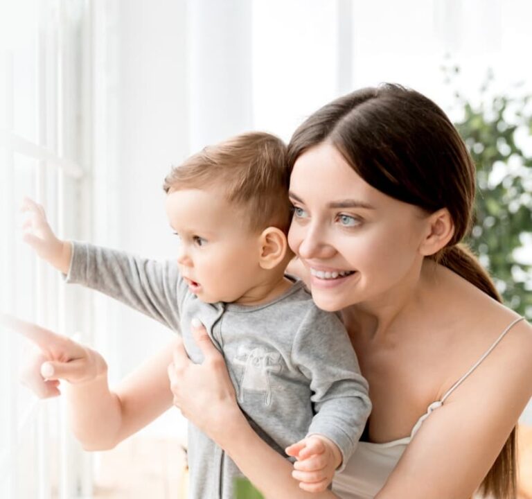 A smiling woman holds a baby, both looking out a window. The baby is pointing outside.