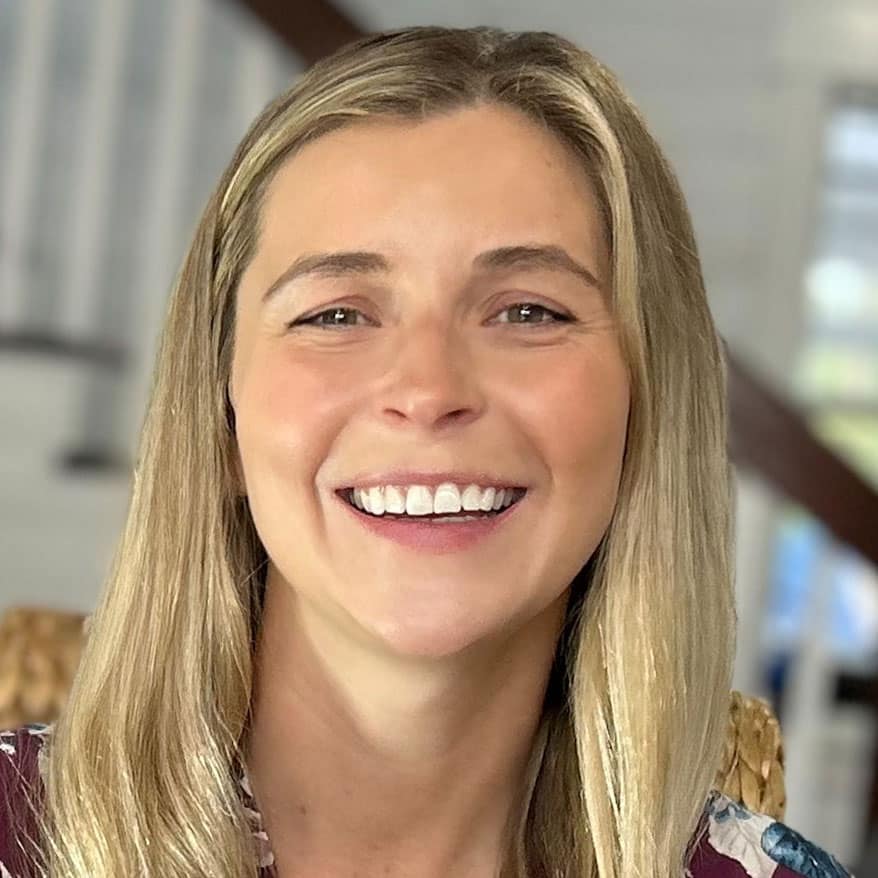 Smiling person with long blonde hair in front of a staircase indoors.