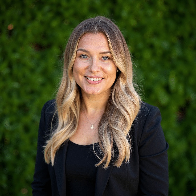 Smiling woman with long wavy hair, wearing a black blazer, stands against a green leafy background.