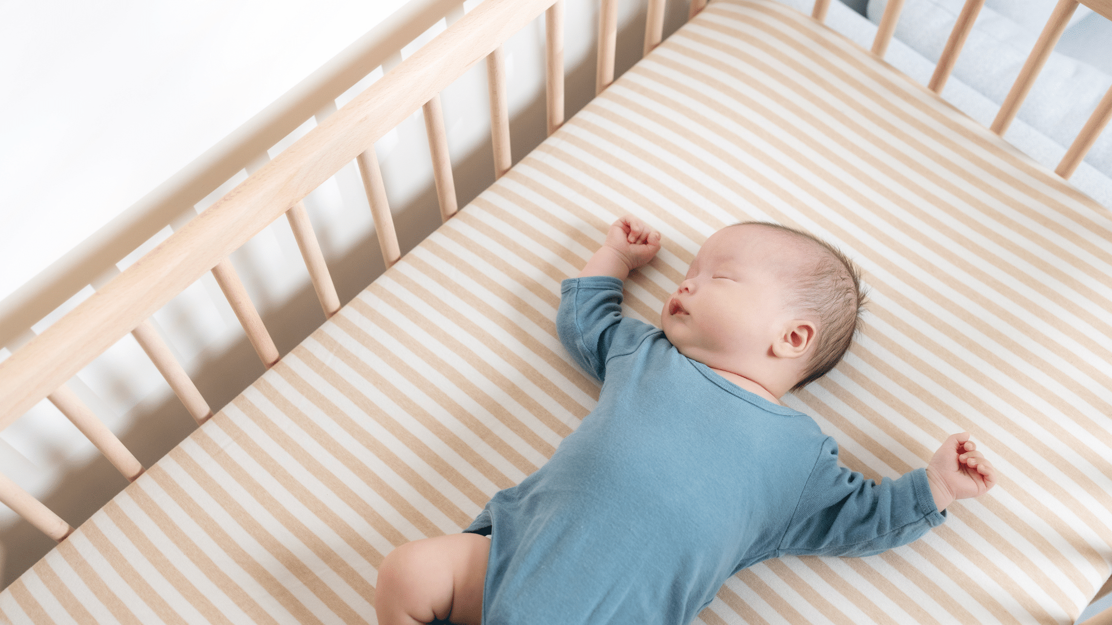 Baby in a blue onesie sleeping on a striped sheet in a wooden crib, arms stretched out.