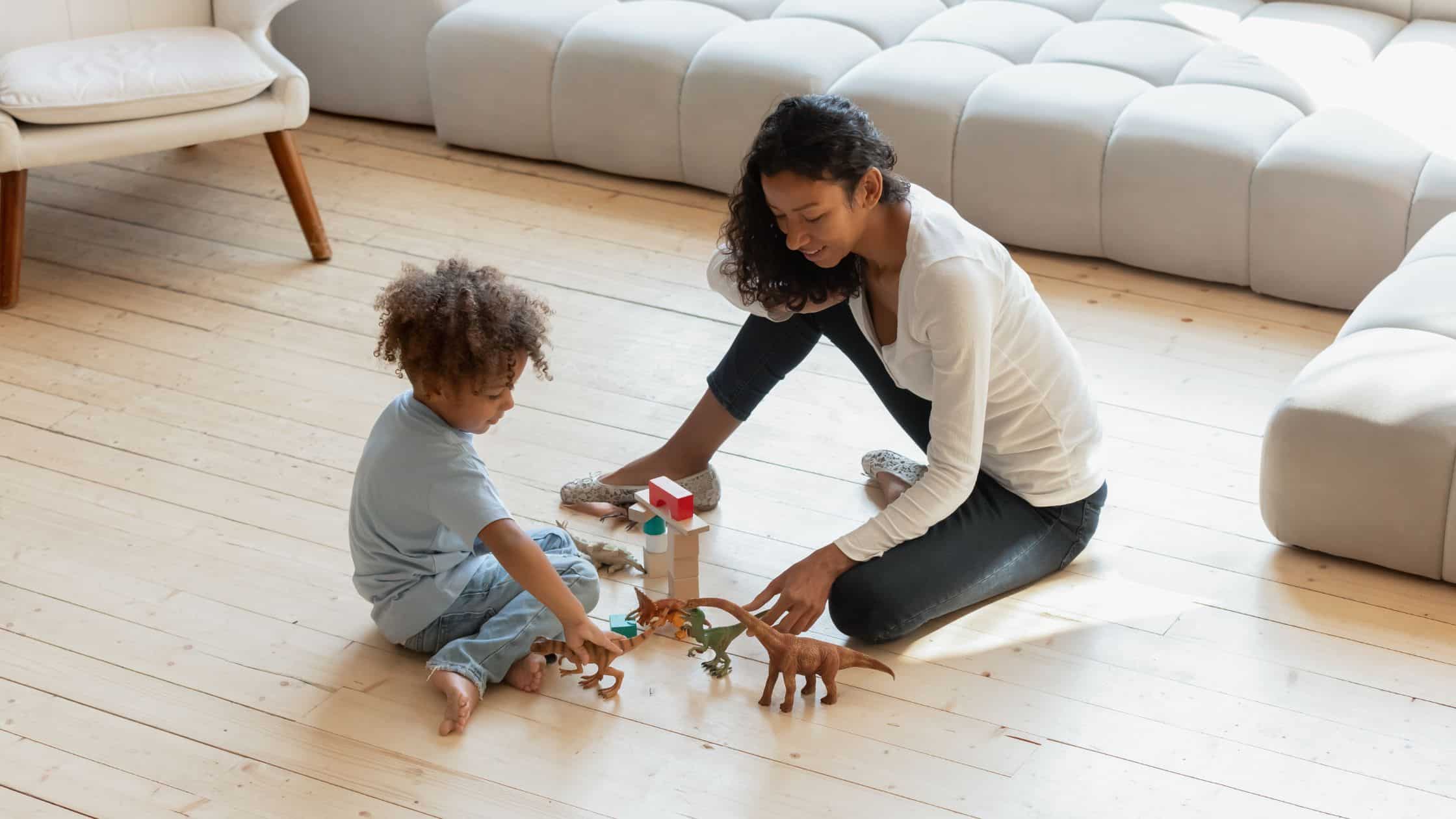 A woman and child sit on the floor playing with toy dinosaurs and blocks in a bright living room.