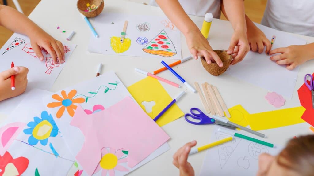Children's hands making colorful drawings and crafts with paper, markers, and scissors on a table.