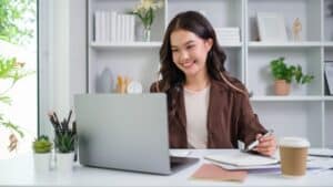 Woman smiling and working on a laptop at a desk with notebooks, pens, and a coffee cup in an office.