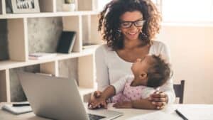 Smiling woman works on a laptop while holding a baby on her lap in a home office, showing the importance of protecting moms’ mental health while balancing work and family.