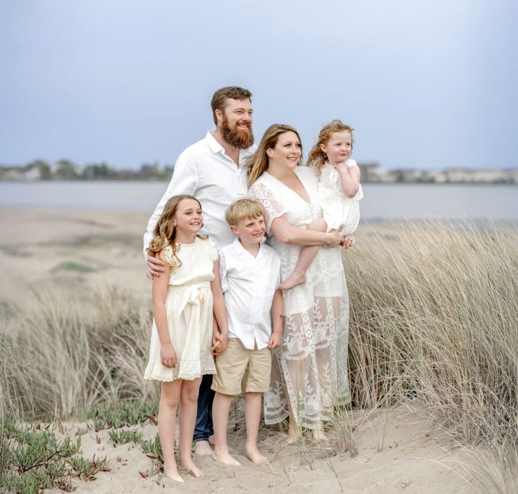A family of five in white clothes stands barefoot on a sandy beach with grass, smiling at the camera, reminiscent of moments shared in the Kat Neill interview.