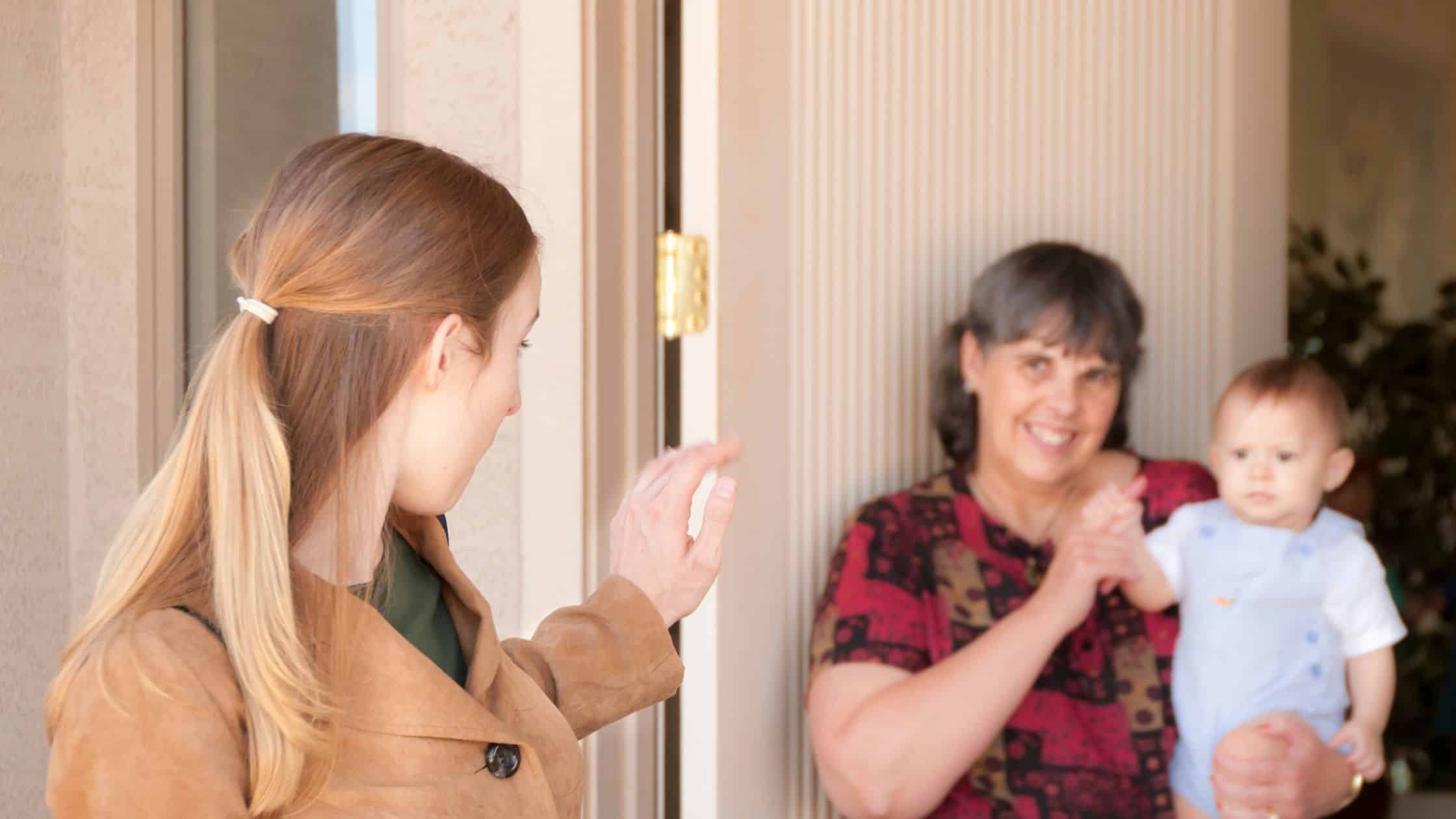 Woman waves goodbye at a door to a smiling older woman holding a baby inside, highlighting the importance of valuing caregiving across generations.