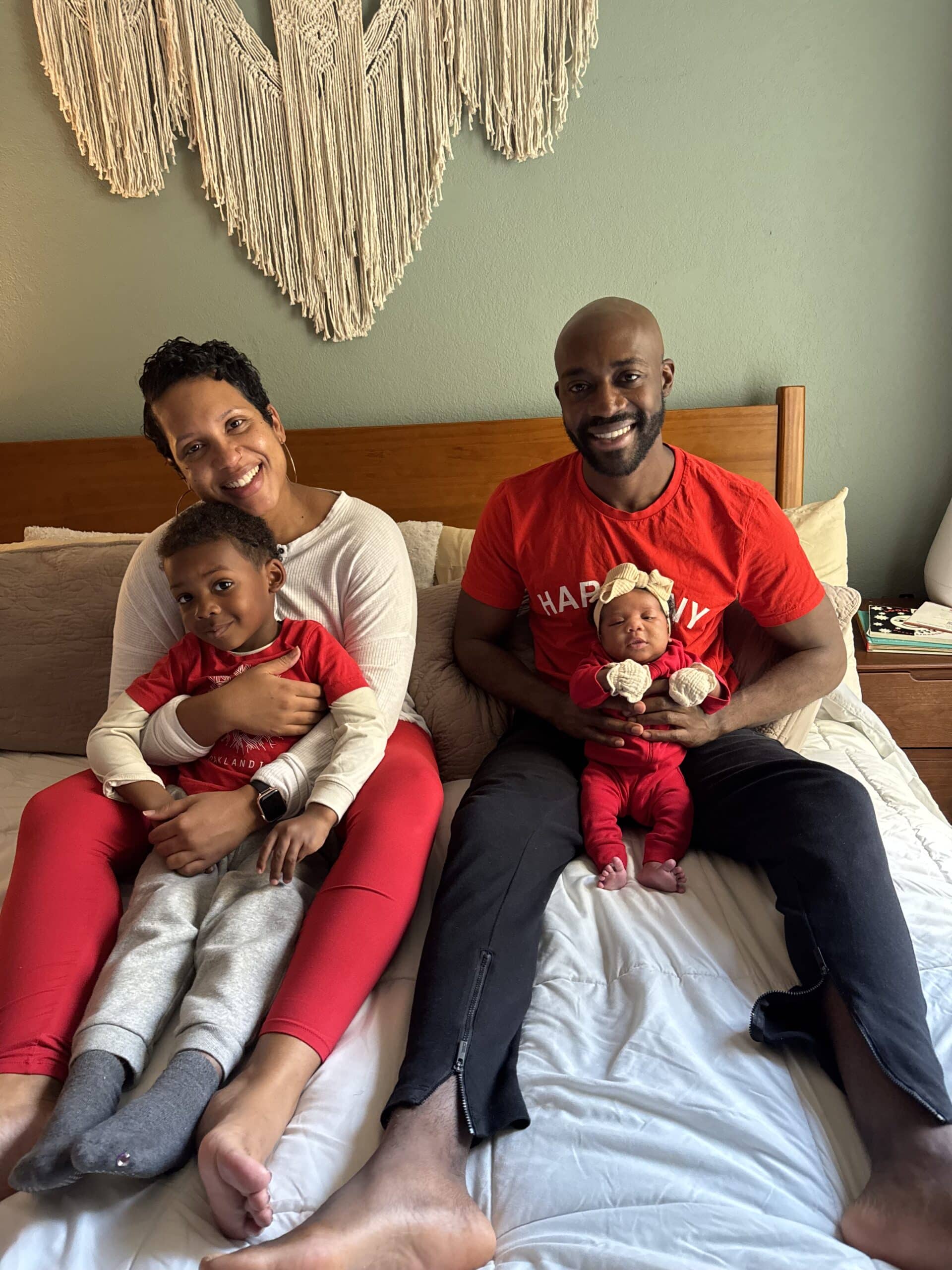 A family of four sits smiling on a bed, parents holding two young children and wearing red and white outfits, in a heartwarming portrait by Katerina Jones.
