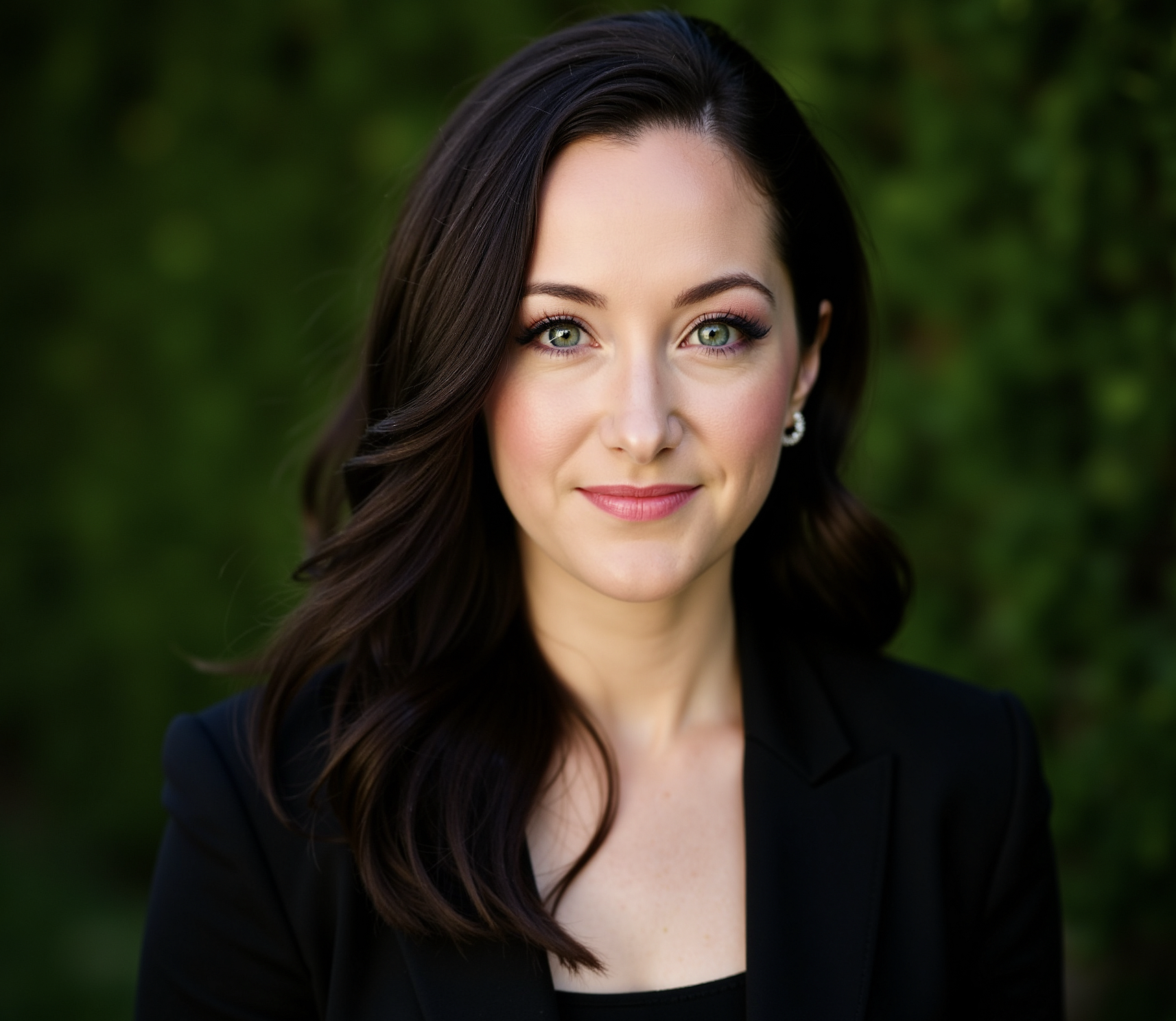 Woman with long brown hair in a black blazer, smiling softly, standing outside with green foliage in background.