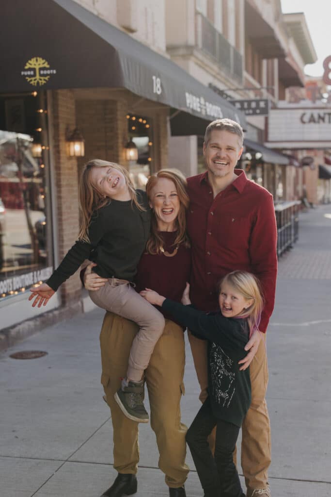 A smiling family of four poses together on a city sidewalk in front of shops and restaurants, reminiscent of a Chelsea Skaggs interview’s warm, candid atmosphere.