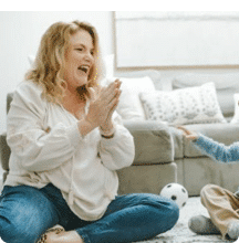 Smiling woman claps while sitting on the floor with a child in a cozy living room.