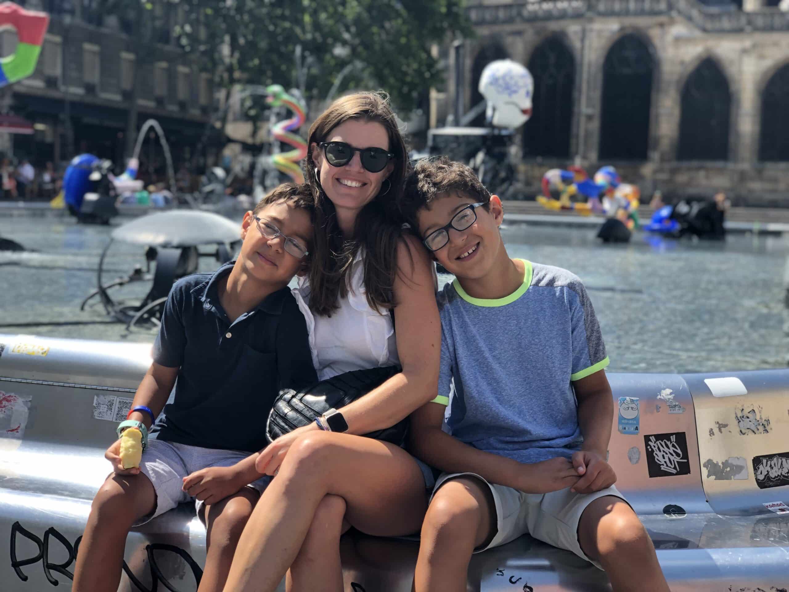 A woman and two boys sit smiling together by a fountain on a sunny day, enjoying a moment that feels straight out of a Kat Neill interview—warm, genuine, and full of connection.