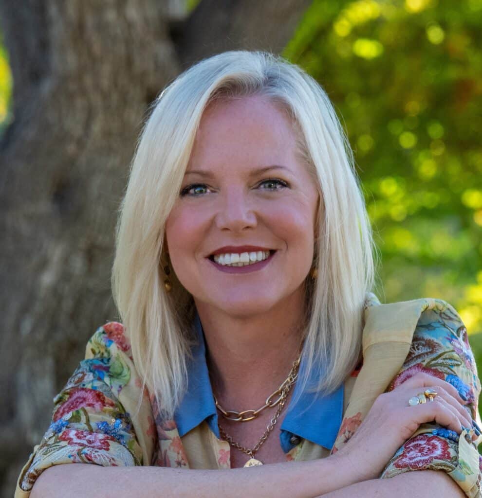 A smiling woman with blonde hair poses outdoors in front of a tree, wearing a floral blouse and gold jewelry.