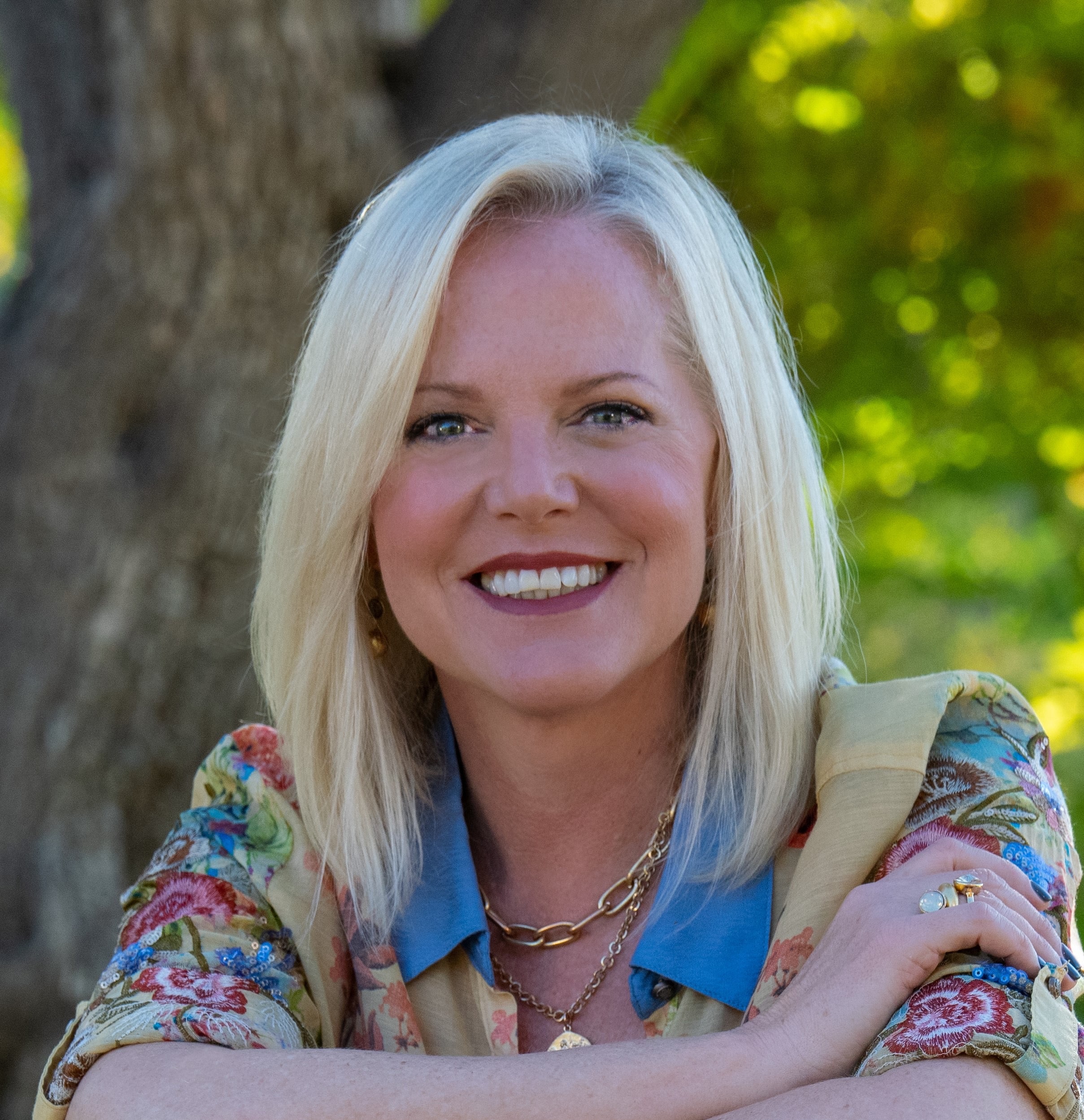A smiling woman with blonde hair poses outdoors in front of a tree, wearing a floral blouse and gold jewelry.