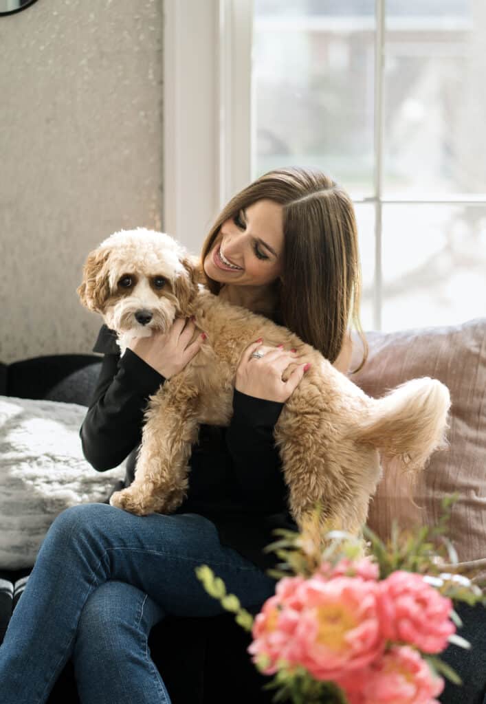 Woman sitting on a couch, smiling and holding a fluffy tan dog, with pink flowers in the foreground—captured during the Lauren Tetenbaum interview.
