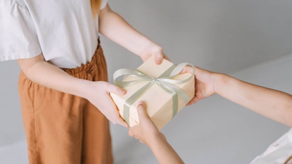 Two people exchange a gift wrapped in beige paper with a light green ribbon against a neutral background.