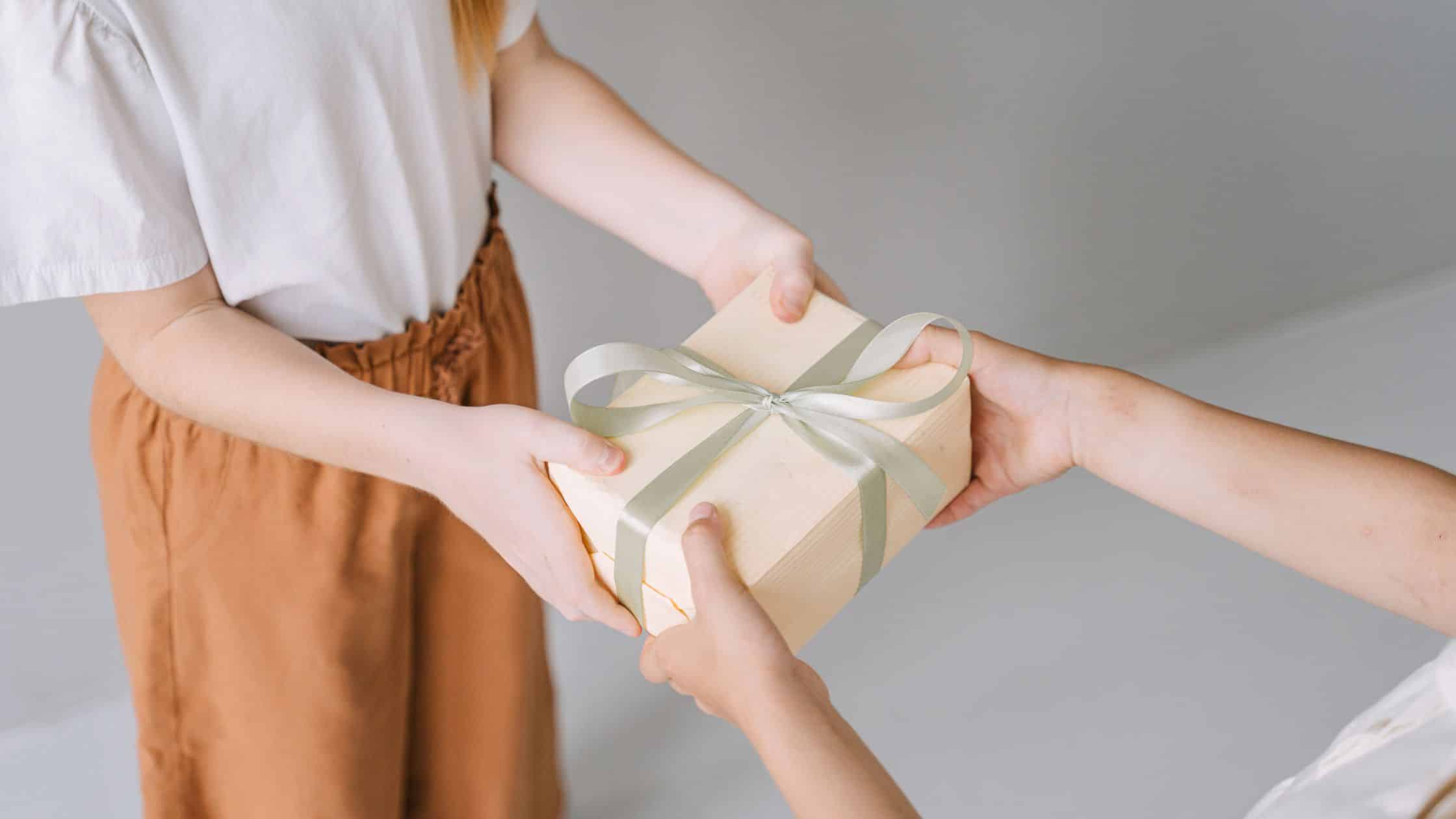 Two people exchange a gift wrapped in beige paper with a light green ribbon against a neutral background.