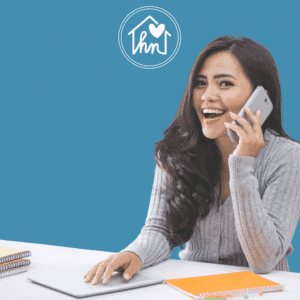 Smiling woman talking on smartphone at desk with laptop and notebooks, blue background with house heart logo, starting her working trial.