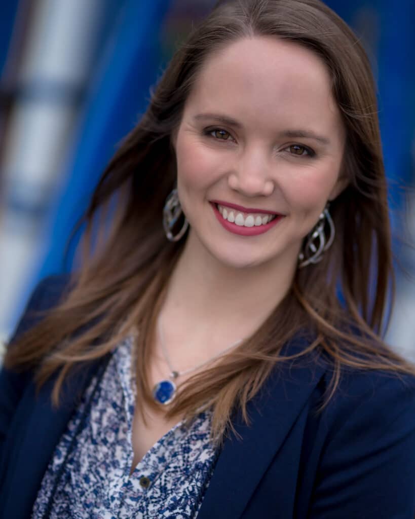 Smiling woman with long brown hair, wearing a blue blazer and patterned shirt, standing outdoors.