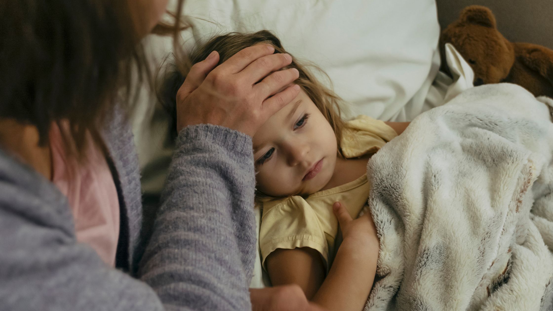 An adult checks the forehead of a sick child lying in bed under a blanket.