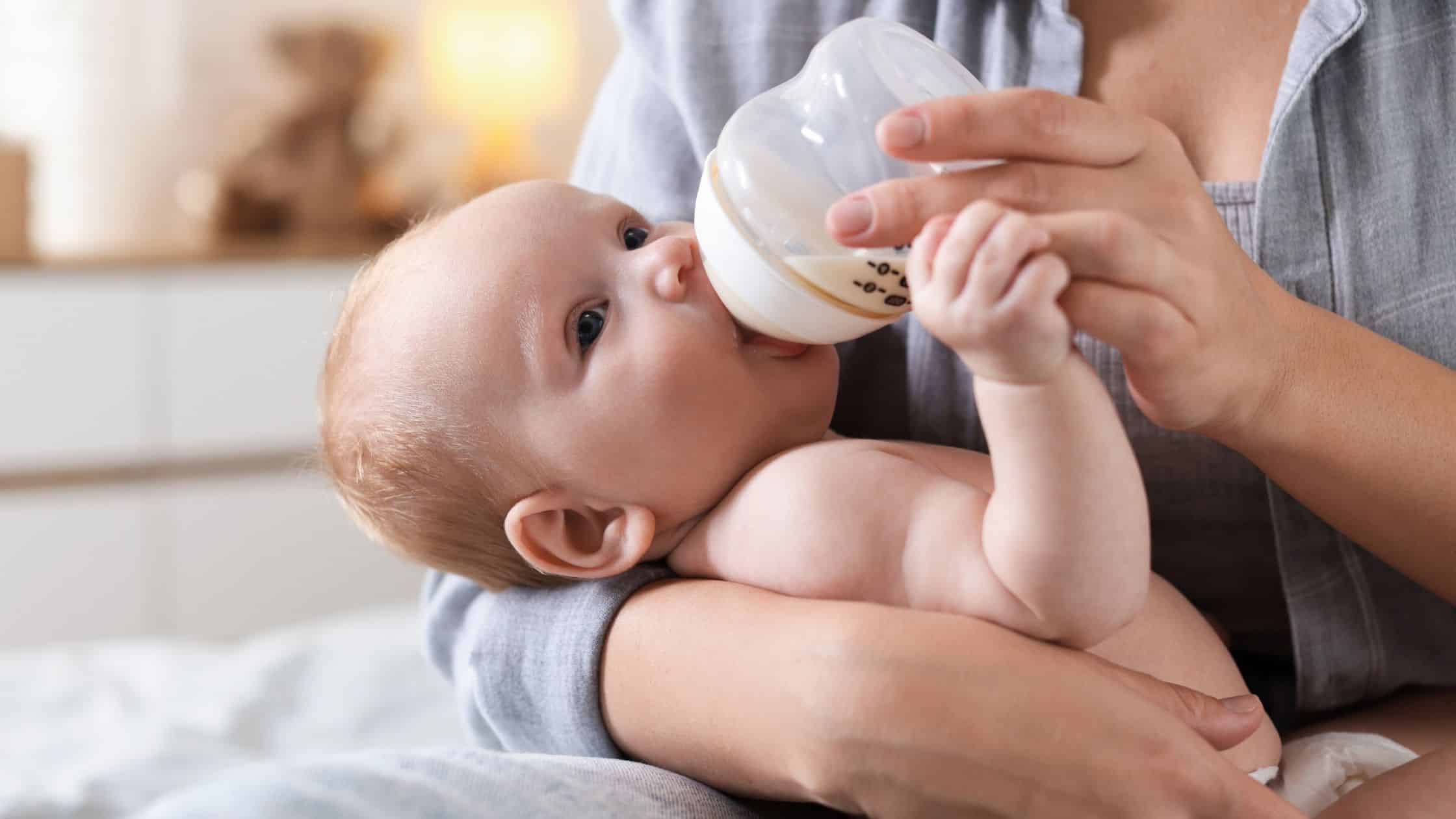 Baby lying in an adult's arms, drinking milk from a bottle, looking up while feeding.