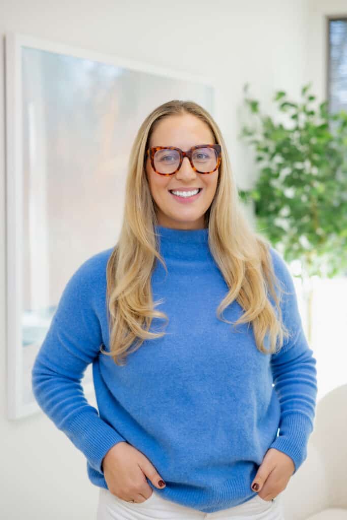 Woman with long blonde hair, wearing glasses and a blue sweater, smiling indoors with a plant in the background. Dr. Chloe Massey