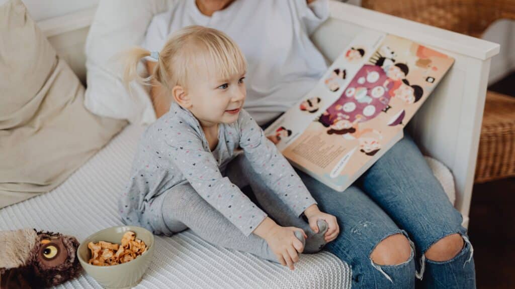 Toddler sits on a couch with a snack bowl; adult next to her holds an open picture book.